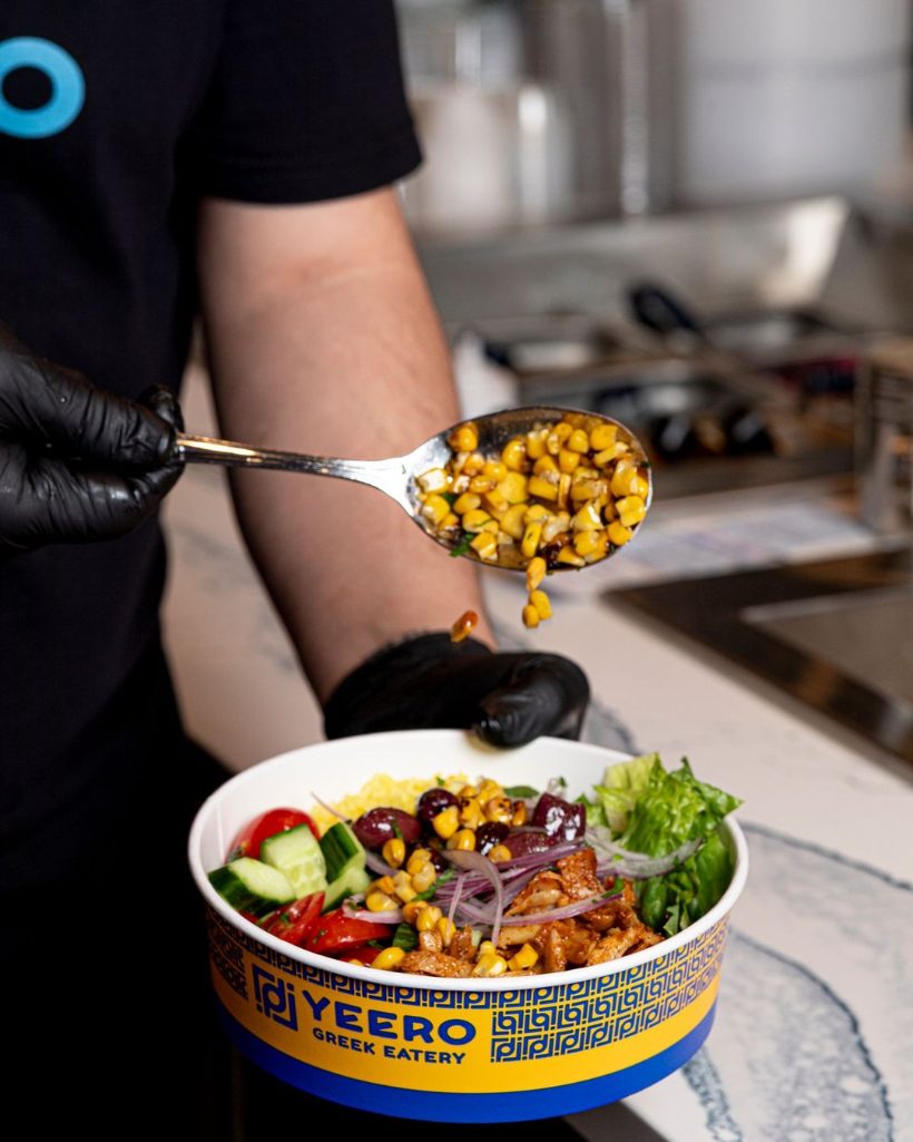 Roasted corn being spooned over a fresh Yeero Greek bowl